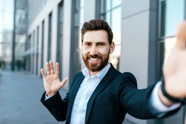 Happy confident handsome european millennial bearded businessman takes selfie, waving hand on street near office building, close up. Successful ceo financial manager, modern blog and online meeting - Photo, Image