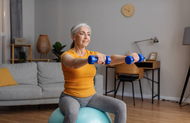 Active retirement. Happy senior woman in sportswear exercising with dumbbells on fitball at home, having domestic training in living room, empty space - Foto, imagen