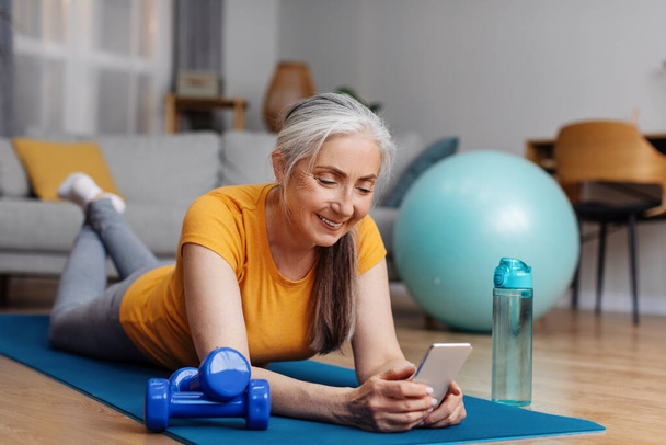 Senior γυναίκα στηρίζεται σε στρώμα γιόγκα, περιήγηση στο διαδίκτυο στο smartphone στο σπίτι, ενώ λαμβάνοντας διάλειμμα μετά το σπίτι fitness, βλέποντας online αθλητικό βίντεο στο κινητό τηλέφωνο - Φωτογραφία, εικόνα