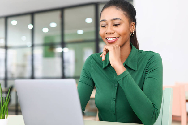 Positive young african-american woman sitting on the desk in the loft style office and using laptop. Cheerful female employee typing on the keyboard, looks at the screen satisfied - Foto, Bild