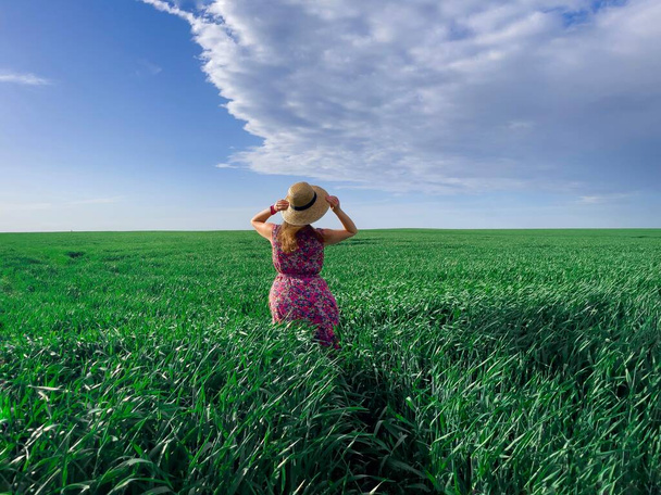 Молодая женщина в платье и соломенной шляпе стоит в поле пшеницы в теплый летний день - Фото, изображение