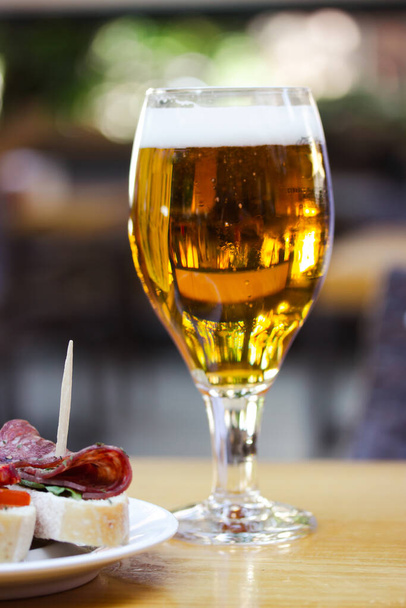 琥珀色のビール。カフェ、バーパブのテラスでは、木製のテーブルの上に黄金の新鮮な醸造小麦ビールとエレガントな食器。伝統的なスペイン料理タパス、ソーセージサンドイッチとアルコール飲料 - 写真・画像