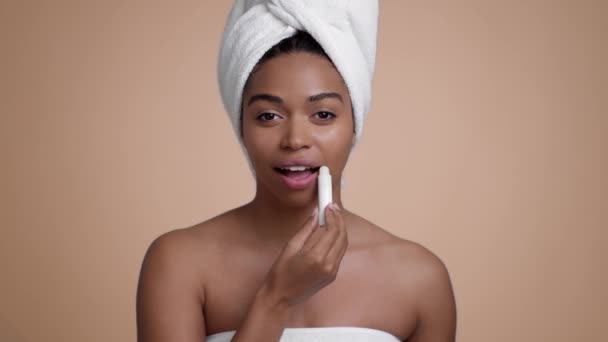 Studio portrait of young pretty african american woman applying hygienic lipstick on lips, posing with towel on head over beige background, smiling to camera, slow motion - Záběry, video