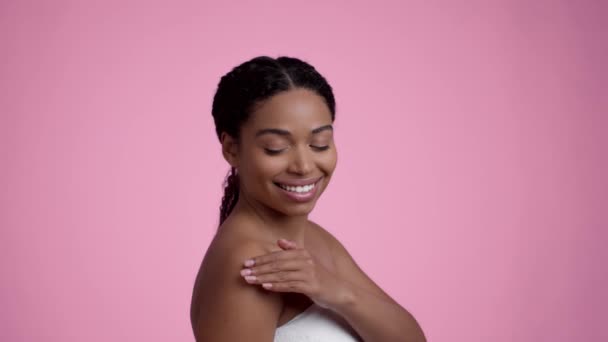 Concepto de mimos corporales. Joven mujer negra bien arreglada envuelta en toalla aplicando crema hidratante en el hombro, sonriendo a la cámara, fondo rosa estudio, cámara lenta - Imágenes, Vídeo