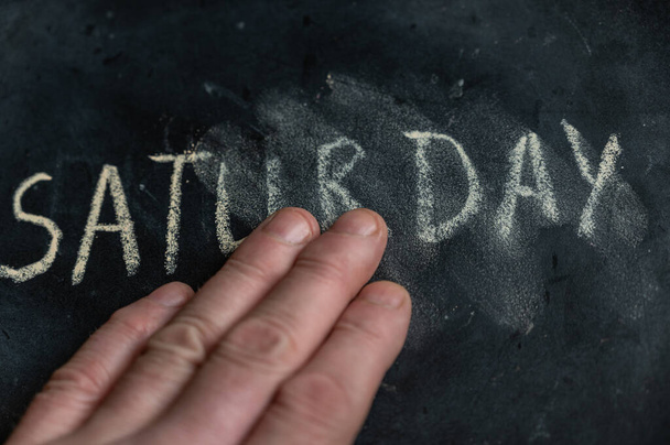 The semi-erased word SATURDAY on the black chalkboard. An adult man's left hand removes the handwritten word with his fingers. Fuzzy letters on black surface. End of calendar month concept - 写真・画像
