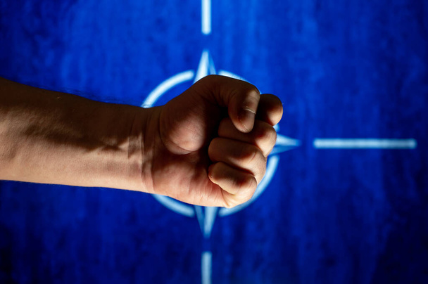 A man's hand clenched into a fist against blue flag emblem. NATO flag is defocused in the background. The firmly clenched fist symbolizes the strength of the alliance. Close-up. Selective focus. - Valokuva, kuva