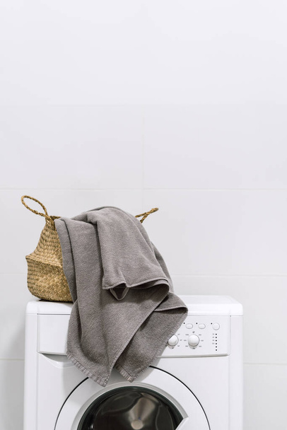 Concepto de limpieza en casa. Vista vertical de la cesta tejida con toalla y lavadero diario de pie en la parte superior de la lavadora moderna, contra el fondo de espacio de copia blanca - Foto, imagen