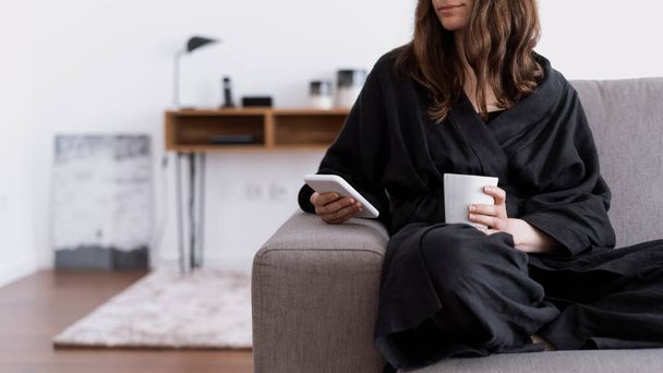 Smiling woman using her modern smartphone, drink beverage and sitting on couch. Cropped view of calm female reading news in online app, hold coffee cup in hand, resting on grey sofa in cozy apartment - Photo, image