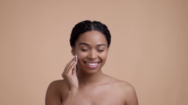 Young beautiful well-groomed black woman cleaning her face with cleansing lotion and cotton pad, smiling to camera over beige studio background, slow motion - Video
