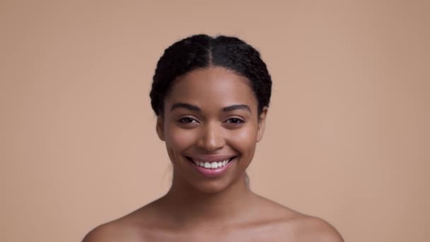 Studio portrait of young pretty african american lady smiling to camera, posing with bare shoulders over beige background, slow motion - Πλάνα, βίντεο