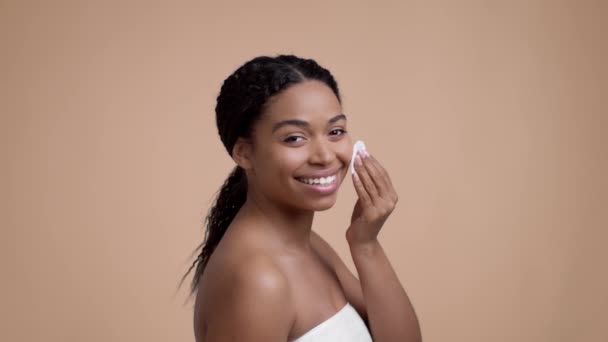 Facial cleansing concept. Young pretty african american lady wrapped in towel cleaning her face with cotton pad, smiling to camera, beige studio background, slow motion - Video