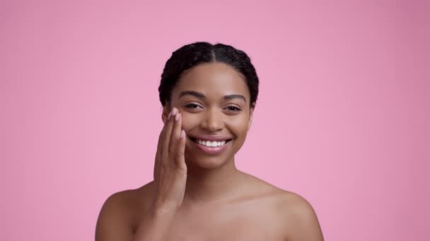Cuidados faciais. Estúdio retrato da jovem mulher americana muito africana aplicando creme de mimos na bochecha, hidratando a pele, sorrindo para a câmera, fundo rosa, câmera lenta - Filmagem, Vídeo
