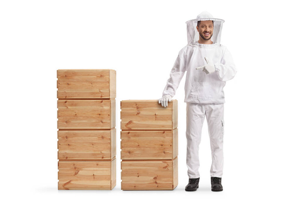 Young male bee keeper in a uniform standing next to wooden crates and pointing isolated on white background - Фото, зображення