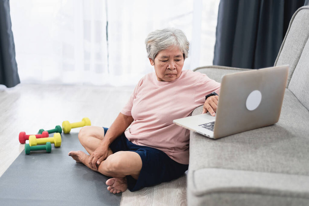 Αρχική έννοια προπόνηση, Ασιατική ηλικιωμένη γυναίκα κοιτάζοντας οδηγό άσκησης στο φορητό υπολογιστή. - Φωτογραφία, εικόνα