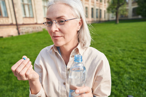 Porträt einer blonden Frau, die eine Flasche Wasser in der Hand hält und Tabletten gegen Kopfschmerzen nimmt - Foto, Bild