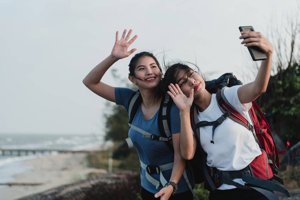 Concepto de turismo al aire libre, dos hermosos mochileros asiáticos tomando selfies y videollamadas usando teléfonos inteligentes. - Foto, Imagen