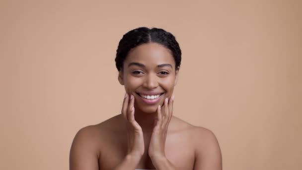 Genç, Afro-Amerikan kadın lenfatik drenaj yüz masajı yapıyor, yüzüne dokunuyor, bej rengi stüdyo arka planında kameraya gülümsüyor, yavaş çekim - Video, Çekim