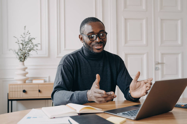 Fiatal jóképű mosolygós afro-amerikai üzletember szemüvegben élvezi a videohívást laptop számítógépen, boldog sötét bőrű férfi alkalmazott beszél kollégájával az online konferencia során - Fotó, kép