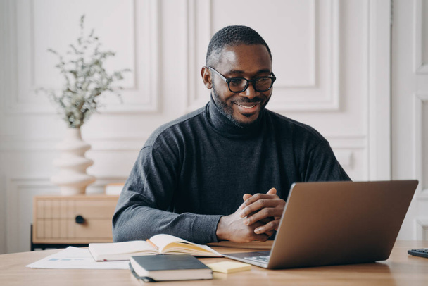 積極的なアフリカ系アメリカ人男性のプライベートコンサルタントは、オンラインセッションを持っていることは笑顔でコンピュータ画面を見ている間、注意深くクライアントに耳を傾け、自宅のオフィスからリモートで働いています。フリーランスのコンセプト - 写真・画像