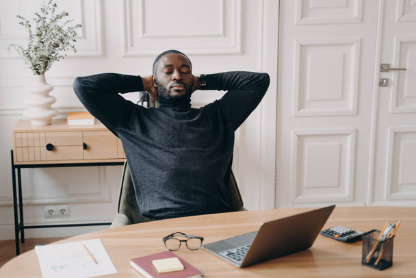 Уставший перегруженный работой афроамериканец-офисный работник с закрытыми глазами и руками за головой расслабляющий на рабочем месте, мужчина-фрилансер, сидящий за столом с ноутбуком и отдыхающий после работы - Фото, изображение