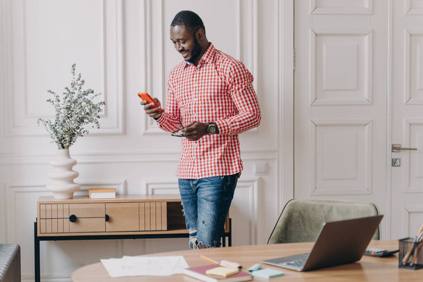 Sebevědomý pohledný africký americký podnikatel pomocí mobilu, stojí v úřadu během pracovního dne, mladý muž tmavé pleti zaměstnanec psaní a dívá se na obrazovku telefonu s úsměvem, chatování on-line - Fotografie, Obrázek