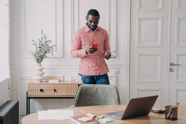 Νεαρός σοβαρός Αφροαμερικάνος επιχειρηματίας με casual ρούχα που στέκεται στο γραφείο με smartphone, χρησιμοποιεί κινητό τηλέφωνο, διαβάζει διαδικτυακές ειδήσεις ή συνομιλεί με πελάτη στο κινητό, κάνει κλήσεις στη δουλειά - Φωτογραφία, εικόνα