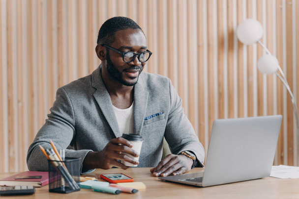 Young smiling african american business in glasses sitting with coffee cup at workplace in office, happy employee looking at computer screen with smile, reading internet news or checking email at work - 写真・画像