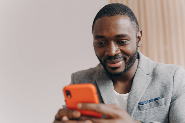 Atractivo sonriente joven africano autónomo en traje formal charlando con los socios en el teléfono inteligente, empresario satisfecho enviando correos electrónicos a la oficina de la empresa antes de la próxima reunión de negocios - Foto, Imagen