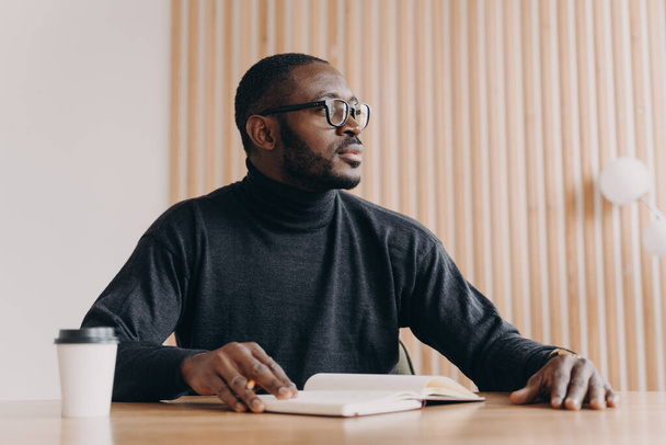 Νεαρός στοχαστικός Αφροαμερικάνος επιχειρηματίας κάθεται στο γραφείο με στυλό και ατζέντα κοιτάζοντας μακριά με σκεπτική έκφραση. Millennial biracial επιχειρηματίας έρχεται με νέες ιδέες στο γραφείο στο σπίτι - Φωτογραφία, εικόνα