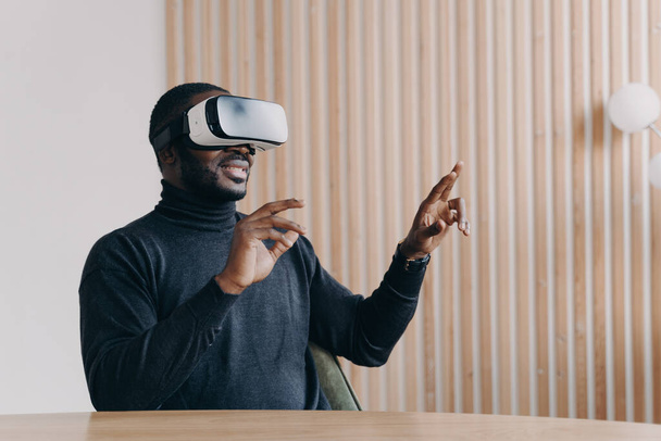 Fröhlicher afrikanischer Mann mit VR-Headset-Brille, der sein Lieblings-3D-Spiel spielt, während er lächelnd am Schreibtisch im Home Office sitzt und mit den Händen nach oben und unten winkt, um Luft zu berühren. Innovatives Technologiekonzept - Foto, Bild