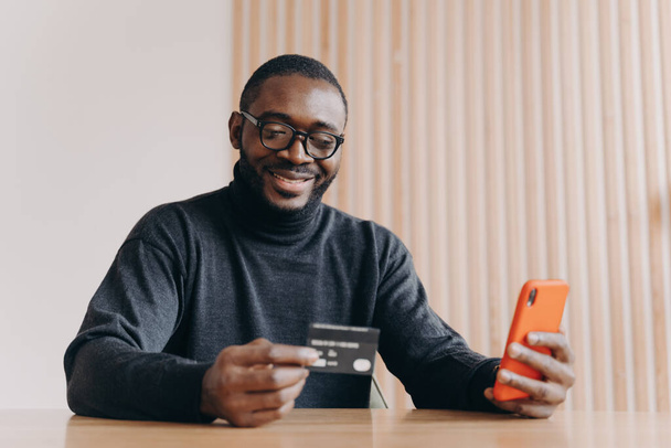 Joyeux entrepreneur afro-américain tenant une carte de crédit et utilisant un smartphone à écran tactile pour faire des achats en ligne tout en faisant des commandes en ligne assis au bureau à la maison. Concept de commerce électronique et de paiement en ligne - Photo, image