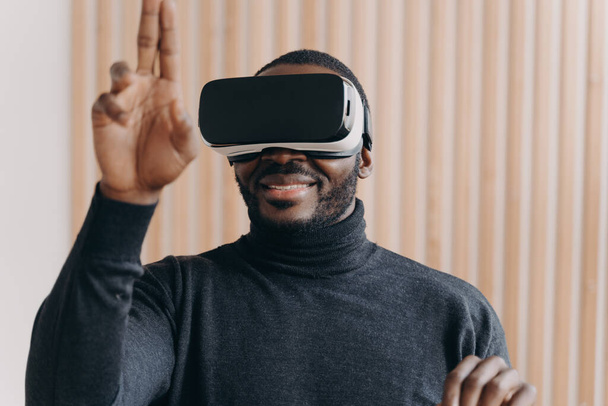 Excited African American man in VR headset glasses enjoying augmented reality while sitting at modern workplace at home, cheerfully raised hand with two fingers up as interacting with cyberspace in 3D - Zdjęcie, obraz