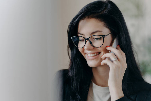 Happy woman lawyer keeps mobile phone near ear, has pleasant conversation with client, discuss working issues, smiles gently, wears transparent glasses, formal wear. Female employee calls someone - Foto, imagen