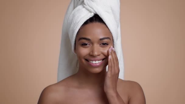 Coccole della pelle. Ritratto ravvicinato di giovane donna afroamericana con asciugamano sulla testa sfregamento crema idratante sul viso dopo la doccia, sfondo beige studio, rallentatore - Filmati, video