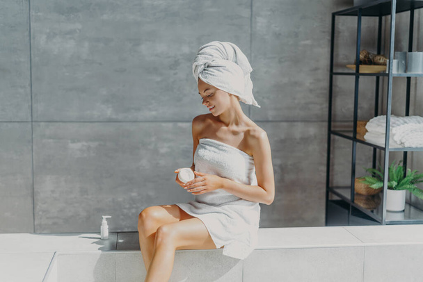 Расслабленная молодая европейская женщина наносит крем-увлажнитель на ноги после принятия ванны, завернутой в полотенце в ванной комнате, наслаждается косметическими процедурами, использует косметический продукт для здоровой кожи. Концепция гигиены - Фото, изображение