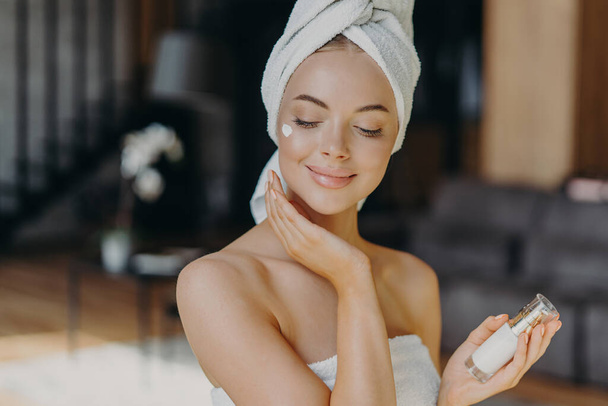 Close up shot of young feminine lady applies face lotion, wears wrapped towel on head, has bare shoulders, smiles with satisfaction, uses cosmetic product. Natural beauty and cosmetology concept - Fotoğraf, Görsel