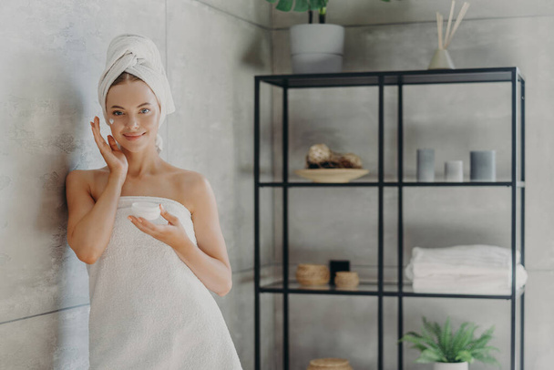 Horizontal shot of young healthy female model uses cream moisturizer, holds jar of cosmetic product, wrapped in towel after showering, stands near wall in cozy bathroom. Skin care procedures - Photo, Image