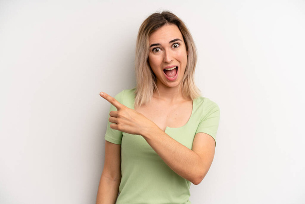 young adult blonde woman looking excited and surprised pointing to the side and upwards to copy space - Foto, afbeelding
