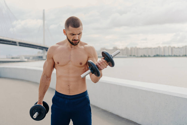 Strong fitness sporty bearded man with thick bristle does exercises with dumbbells, pumps up biceps, poses outdoor, has naked torso, breathes fresh air. Fitness, sport and lifestyle concept. - Photo, Image