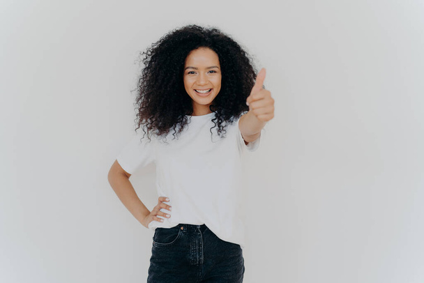 Photo of happy curly woman with crisp hair raises thumb up, gives approval, says sounds good, makes supportive gesture, keeps other hand on waist, dressed casually, isolated over white background - Photo, Image