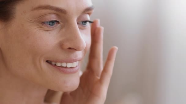 Anti aging skin care. Close up portrait of beautiful middle aged woman with wrinkles applying caring cream on face, touching her skin, tracking shot, slow motion, empty space - Footage, Video