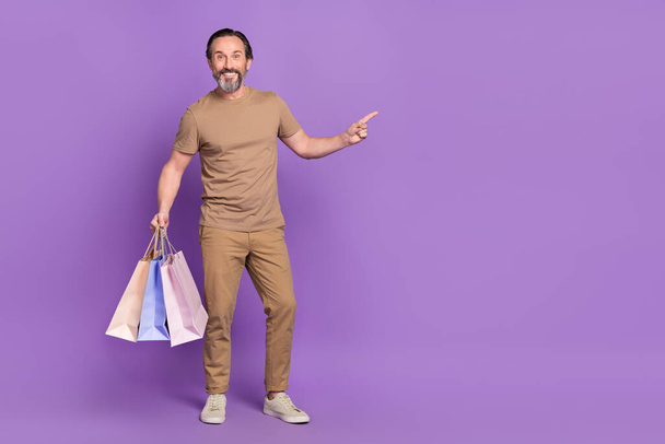 Full length photo of mature nice man hold bags index promo wear t-shirt trousers sneakers isolated on purple background. - Foto, afbeelding