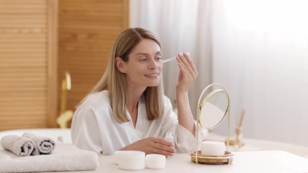 Gezichtsverzorging. Gelukkig middelbare leeftijd vrouw toepassen zorgzame serum op het gezicht, kijken naar spiegel naar de badkamer en glimlachen, slow motion, lege ruimte - Video