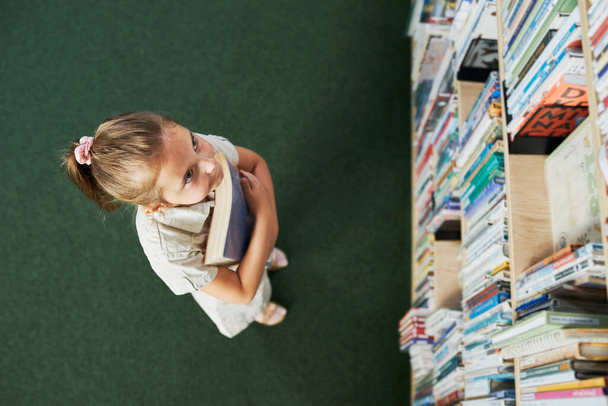 Sttudent looking at top bookshelf in school library. Smart girl selecting literature for reading. Books on shelves in bookstore. Learning from books. Back to school. Elementary education. Benefits of everyday reading. Curious child - Фото, зображення