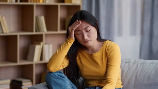 Migréna. Vnitřní portrét mladé asijské ženy trpící akutní bolestí hlavy, dotýkající se jejího bolestivého čela, sedící doma, zpomalený pohyb, volný prostor - Záběry, video