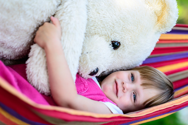 Little cute girl lying on hammock with large teddy bear - Photo, Image