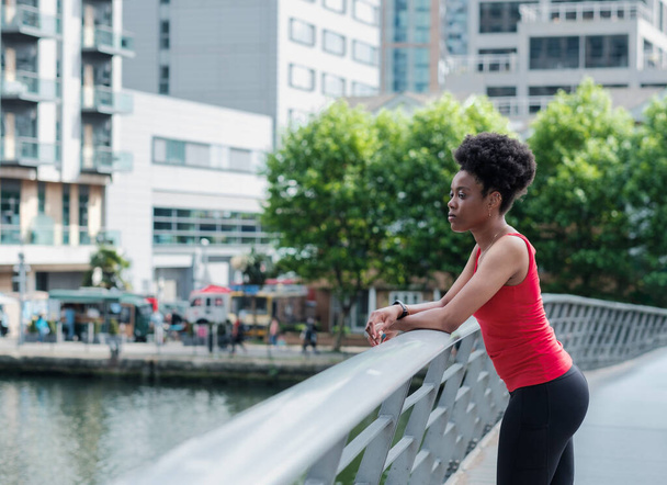 A black sport young woman is looking away leaning on a banister bridge. Her hair is black and curly. She is wearing black leggings and sleeveless red t-shirt. Modern buildings on the background. - Foto, imagen