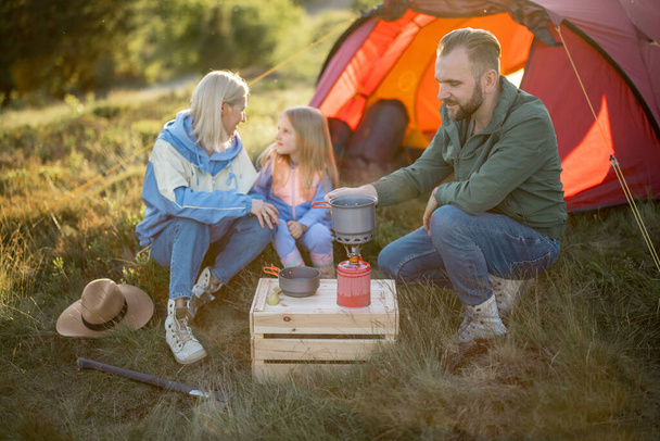 Paar mit kleinem Mädchen picknicken auf dem Campingplatz, während sie mit Zelt in den Bergen unterwegs sind. Junge Familie verbringt Urlaub gerne in der Natur - Foto, Bild
