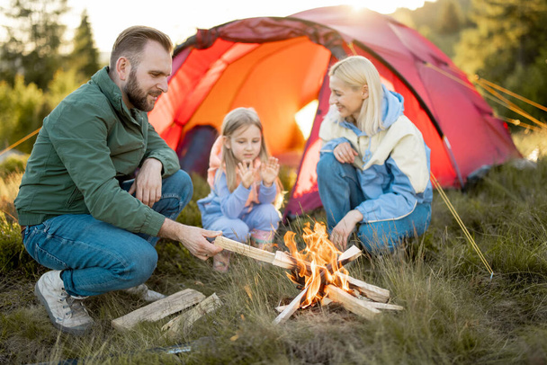 Young adult couple sit with a little girl by the campfire at campsite, traveling with tent on nature. Family spend summer time hiking in the mountains - Photo, Image