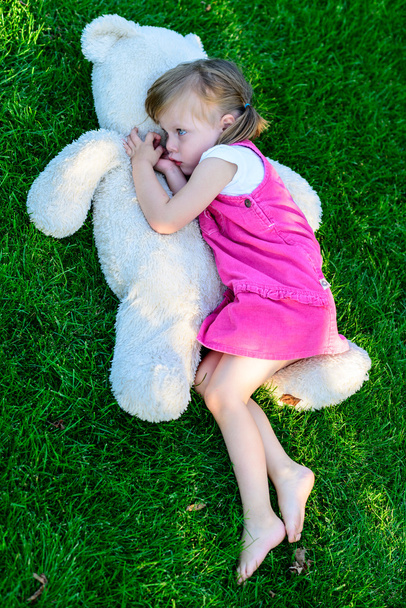 Sad little girl lying on grass with large teddy bear - Foto, afbeelding